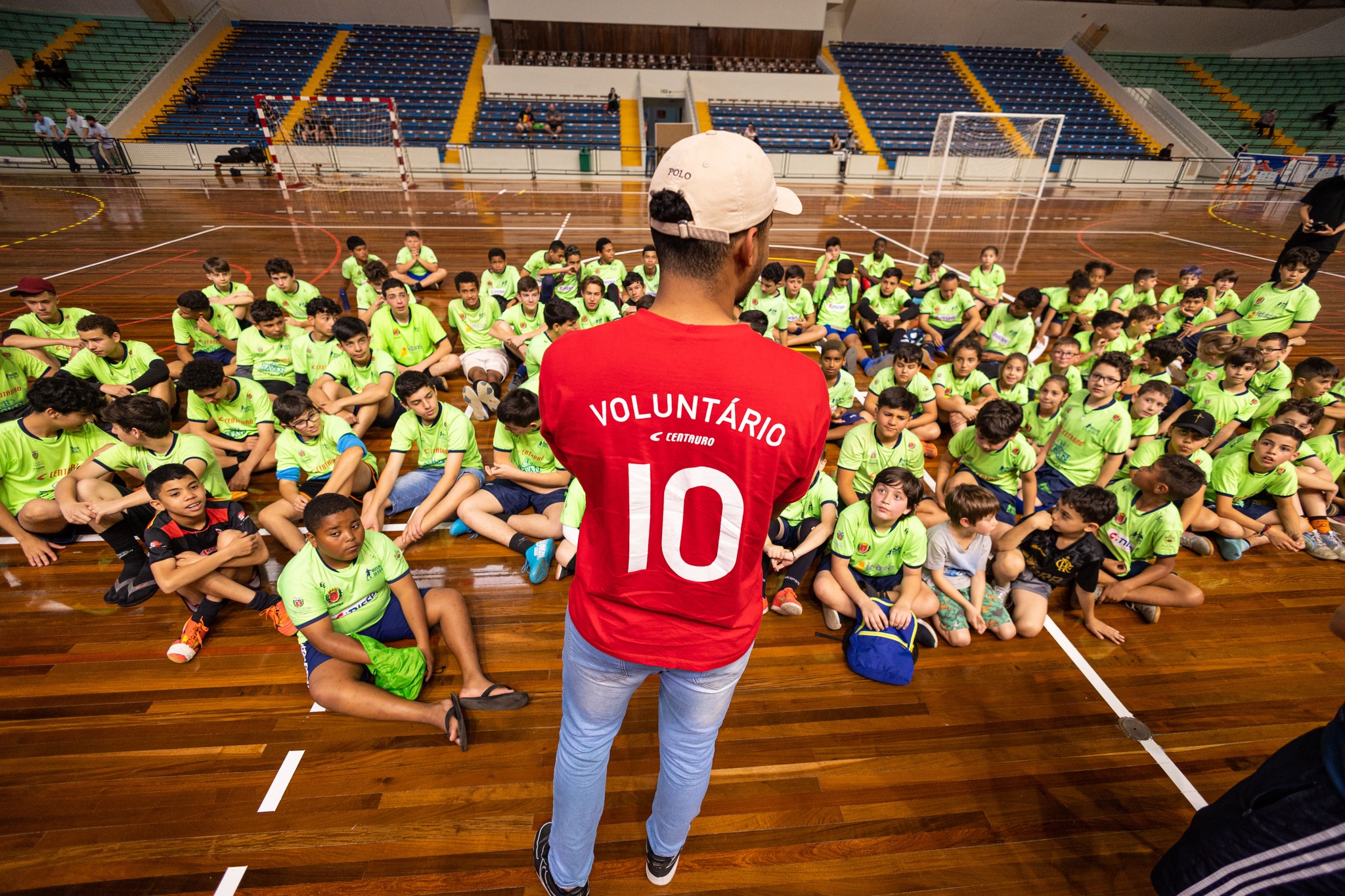 a foto mostra várias crianças atletas sentadas ouvindo um dos técnicos voluntários falar