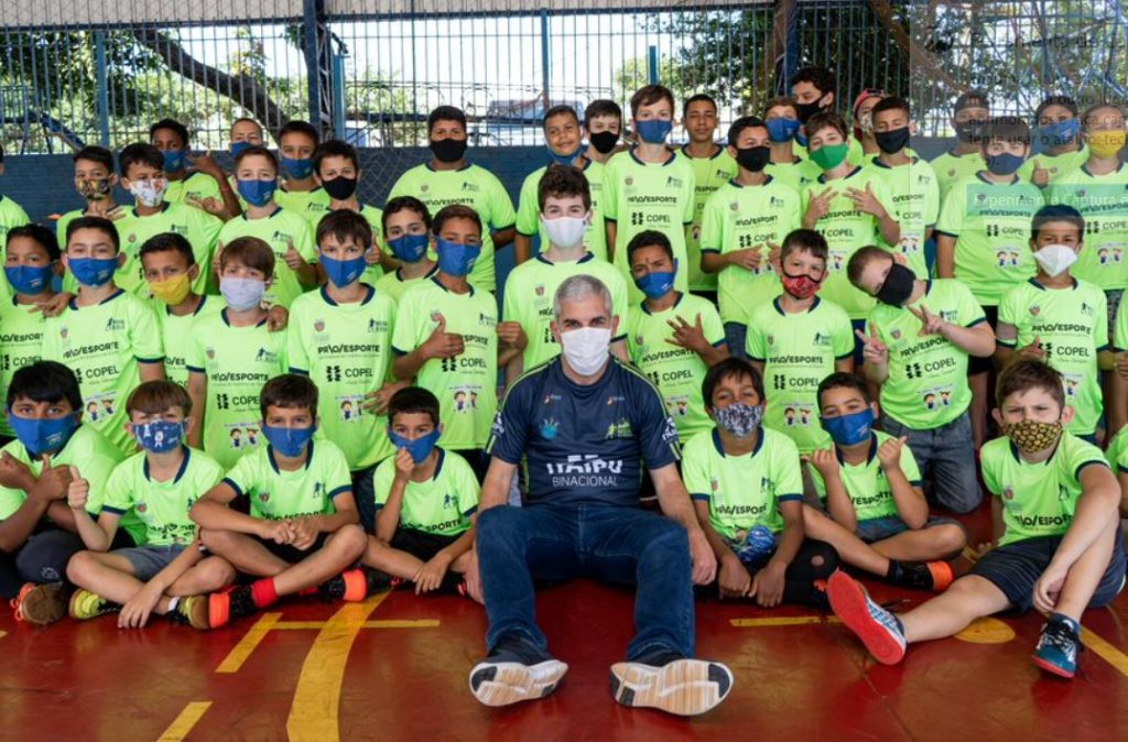 foto mostra o jogador Ricardinho sentado numa quadra trajando roupa azul marinho. Ao seu redor, várias crianças estão sentadas e estão vestindo a camisa do projeto de cor amarelo limão. As crianças estão posando para a foto. Todos estão usando máscaras faciais para prevenção do covid-19.